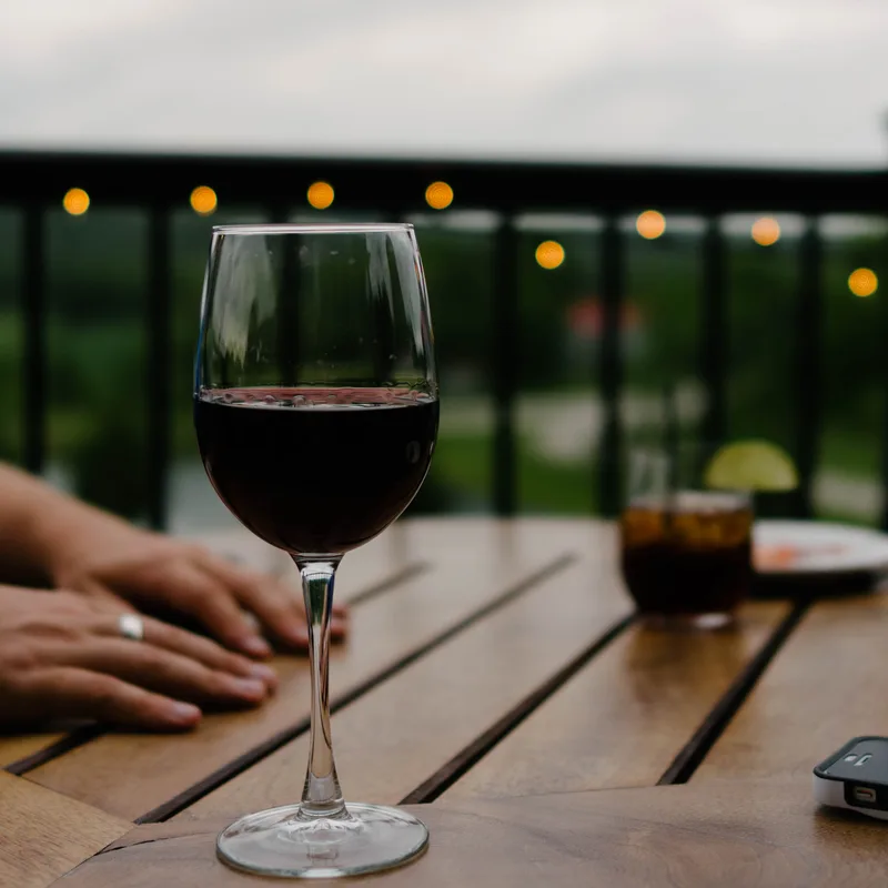 A glass of red wine on a wooden table on a terrace at dusk, with festoon lights strung along the railing behind